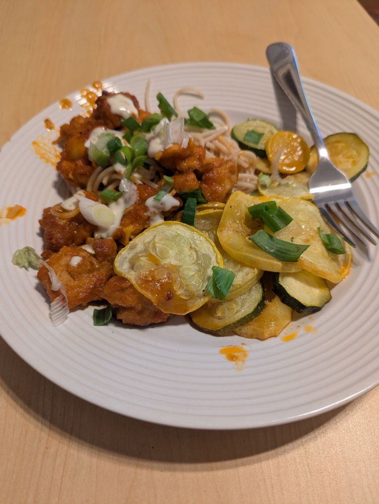 Browned and breaded chicken with buffalo sauce next to roasted yellow and green zucchini and squash. Topped with ranch and green onion slices on a bed of brown rice noodles, sitting on a white plate with a fork on a wooden table
