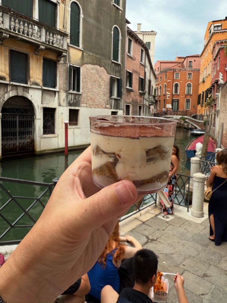 A pot of Tiramisu being held on the air by a hand with a canal and some old Venetian buildings in the background.