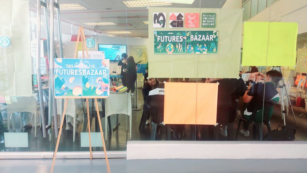An easel board with a colorful sign This Way to the Futures Bazaar in front of a glass wall with two posters of the same sign and 4 logos.