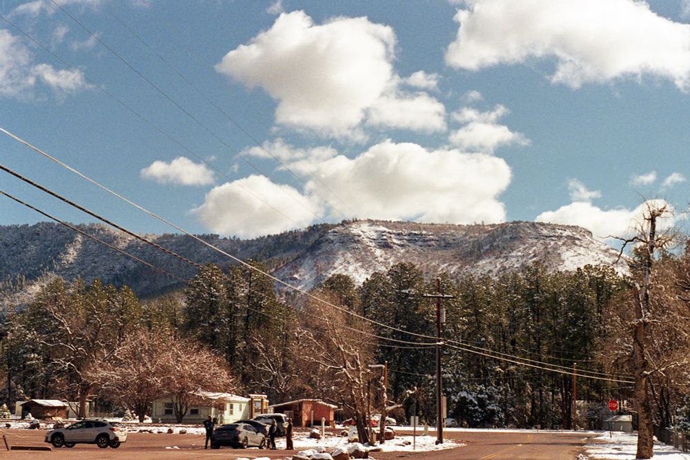 A landscape shot of snow dusted mountains. There are fluffy white clouds on a light blue sky. The mid ground is trees that stretch across the horizon and are lightly dusted with snow. There are small buildings seen on the bottom left and some cars and people in a parking lot. 