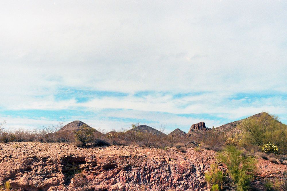 A shear rock outcropping is in the foreground. The rock is tan and slightly red and there is a green bush growing in the bottom right. In the background some mountains can be seen. The sky is mostly wispy clouds and some light blue sky can be seen through them. Some of the bushes have small yellow flowers to the right of the image. 