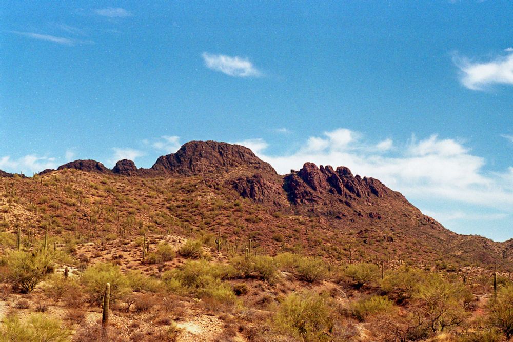 A mountain along Vulture Mine Road in Arizona. There is some green desert brush and a few saguaros in the foreground. The mountain is centered in the shot and the sky is light blue with some wispy white clouds behind and to the right of the mountain. The mountain has three sections: the left slopes in two small peaks; the middle the tallest peak, plateaued at the top; and the right many more jagged sections of the peak. 