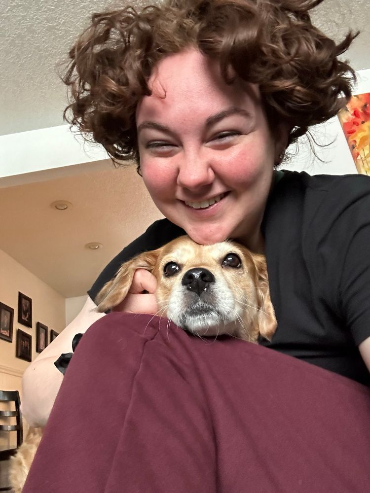 Me with short hair at my ears, a long pixie I guess, sitting in my living room wearing a black shirt and burgundy sweats, cuddling Annie (a spaniel mix who looks like a small golden retriever) who is staring off into the middle distance