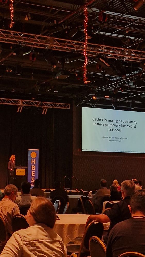 Middle-aged white woman standing by podium and HBES flag in large event space under screen with title "8 rules for managing patriarchy in the evolutionary behavioral sciences" and audience members in front. 