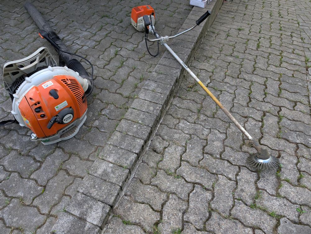 Stihl FS 160 mit Unkrautbürste und Stihl Laubbläser nebeneinander auf einem gepflasterten Hof 