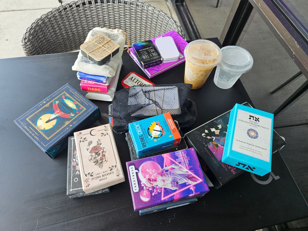 A collection of tarot decks and other divination tools. Accompanying them are a cup of iced coffee, a cup of water, and the book Pronoun Trouble by John McWhorter.