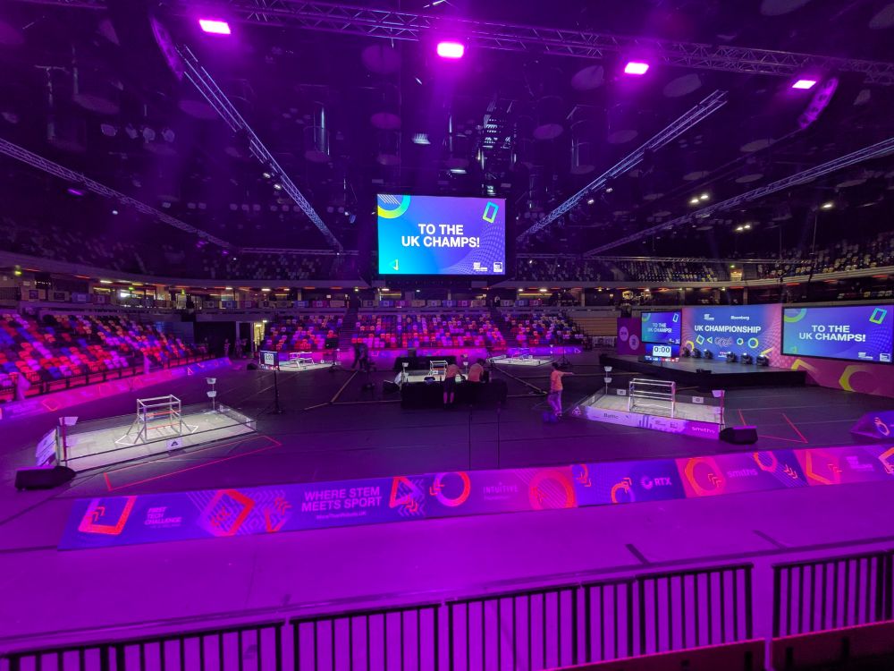 An arena with purple lighting, and five game fields, surrounded by sponsor hoarding and tiered seating. A jumbotron style screen hangs above the field. 
