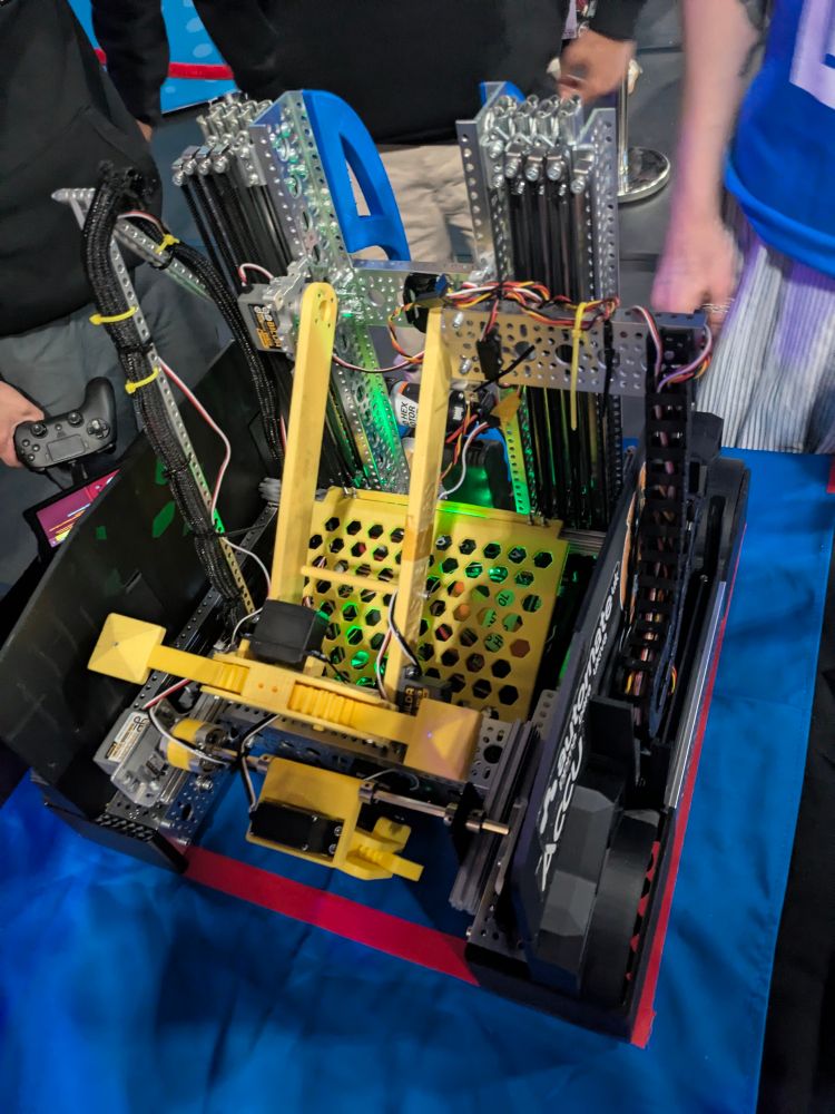 A bright yellow robot with green LEDs, and two arm mechanisms, one on an elevator and another horizontally expanding. 