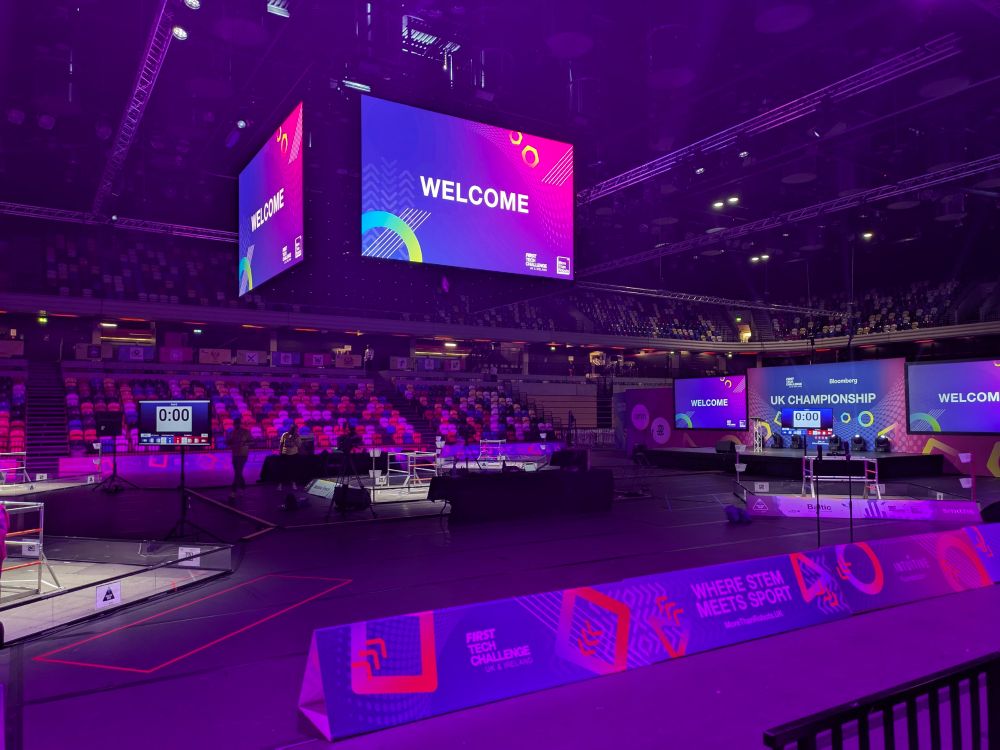 An arena with purple lighting, and five game fields, surrounded by sponsor hoarding and tiered seating. A jumbotron style screen hangs above the field. 