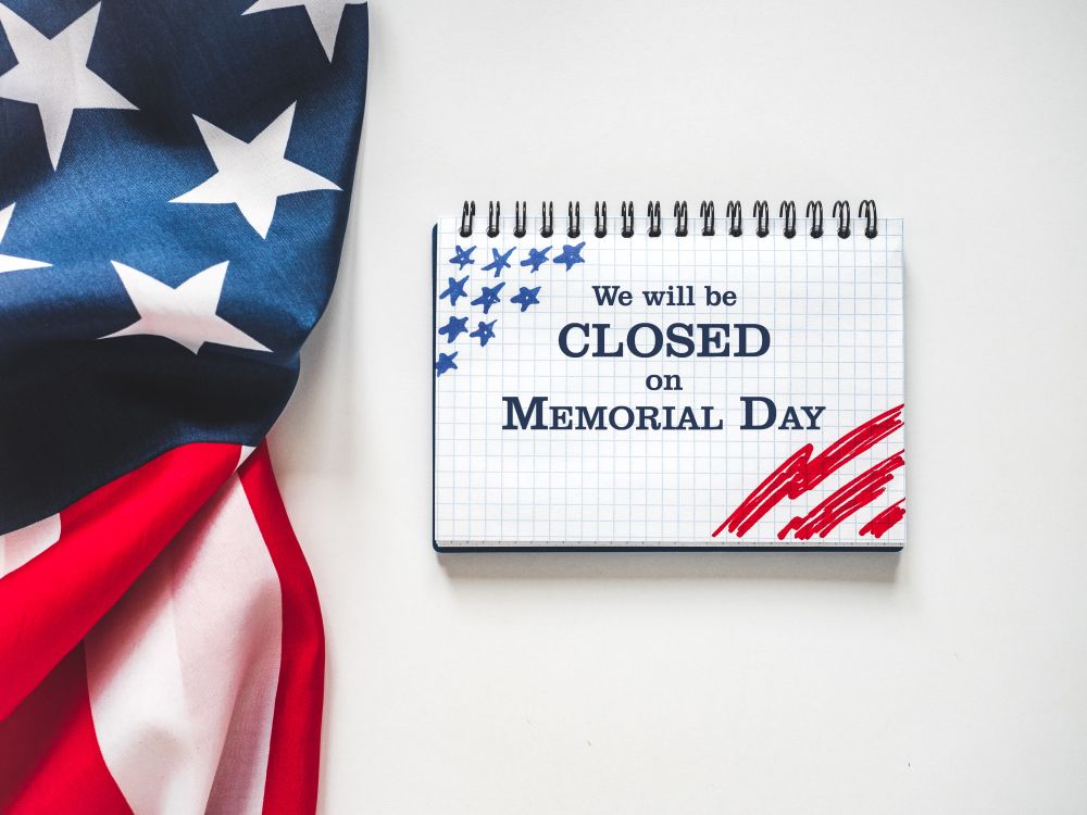 Image of an open notebook with the words "We will be closed on Memorial Day" written on its page. A folded American flag is pictured to the left of the open notebook.