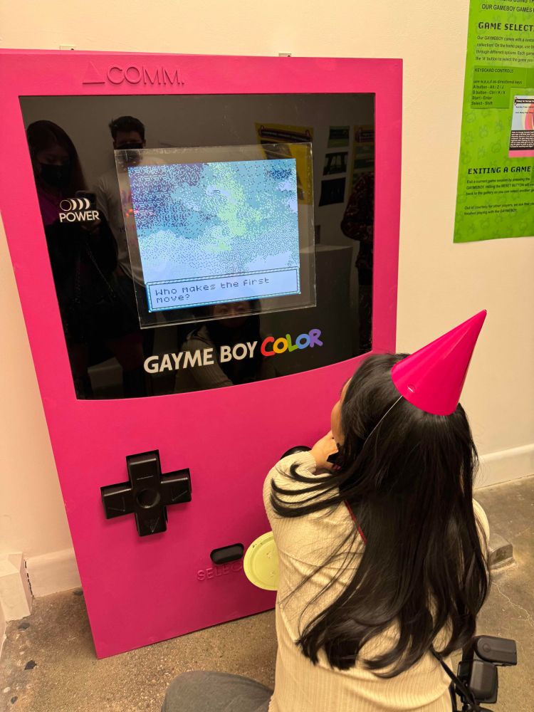 a photograph of a girl with a bright pink party hat playing a game on a giant, five-foot tall gameboy. The gameboy's marquee reads "Gayme Boy Color"