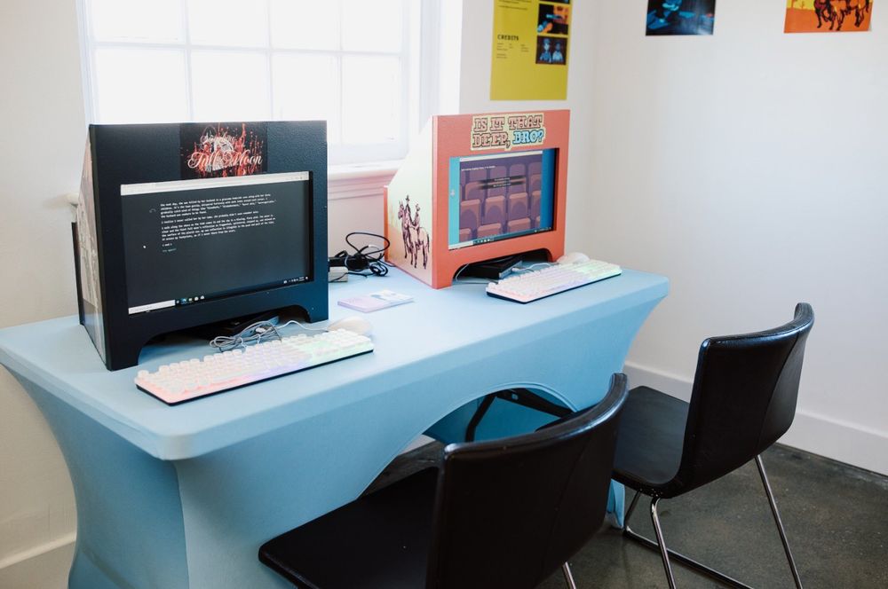 Two custom-made PC cabinets rest on a table with a light-blue table cloth. The PC Cabinet on the left is black and features a browser-based game called "Searching for the Full Moon". The cabinet on the right is bright orange and features a game called "is it that deep, bro"