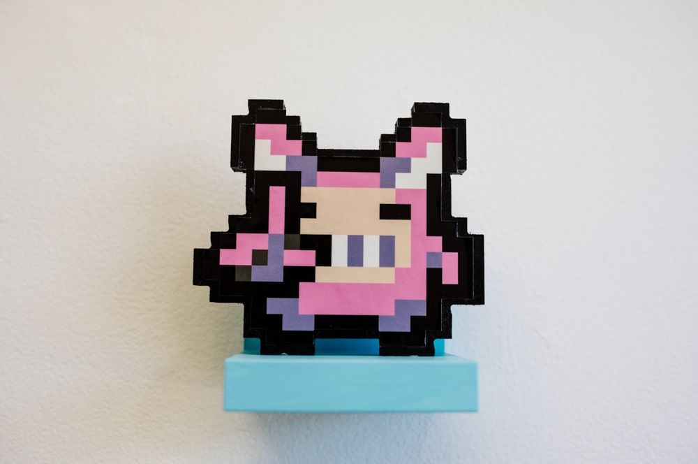 a photo of a pixelated flat figurine of FREEPLAY's mascot, Buggy! Buggy is an 8-bit funny, bunny-like gremlin creature who is pink. He is grinning mischievously, posing with the "L" sign on his right hand.