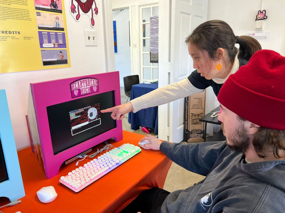 A photo of two people playing a game by Alec Robbins called "Heartbreak High". The game is hosted on a custom-made cabinet, which is hot pink. The game's logo is featured on the marquee.