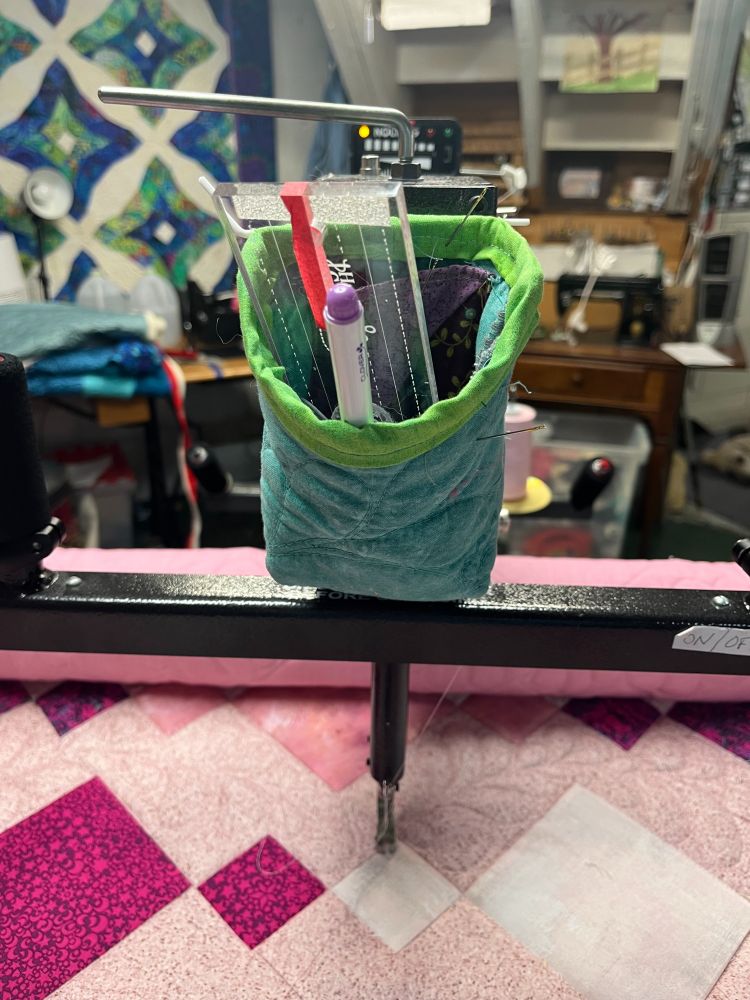 The front of a Nolting Longarm quilting machine. A pink and white quilt is at the bottom of the photo as it is on the frame being quilted. There is a green notions bag attached to the post of the machine in front in which sits a pen and my favorite ruler with red tape on it. You can't see the snips. 