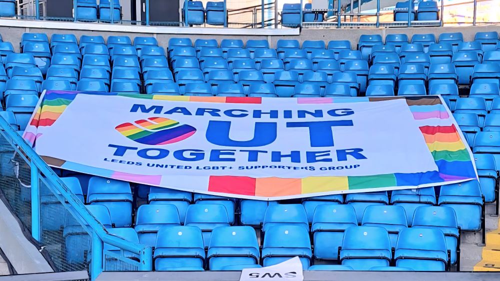 Large banner with M-Out name & rainbow-smiley logo, now edged in Progress Pride colours, stretched over blue seats in SW corner