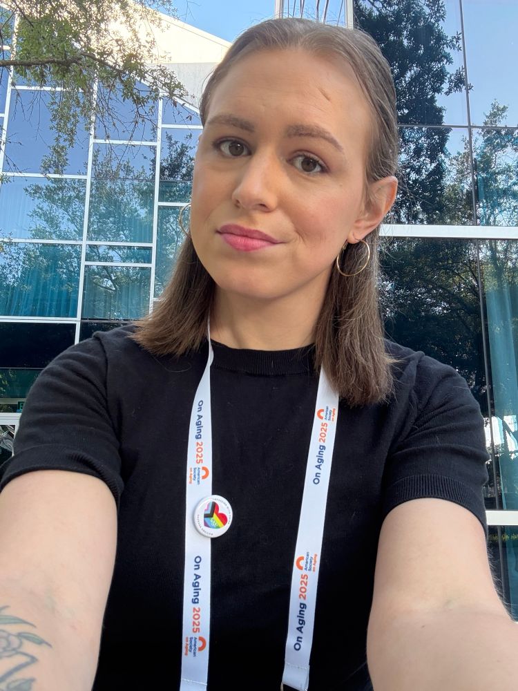 A photo of Jane, a woman with shoulder length light brown hair, in a black short sleeve sweater. Jane is in front of a building with square glass windows. 
