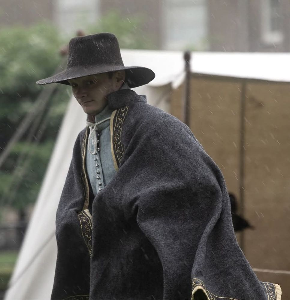 An image of an English Civil War reenactor dressed in a wide-brimmed, rain-flecked brown hat, gold-trimmed cloak and blue doublet. Photo by Yorkshire Pictures