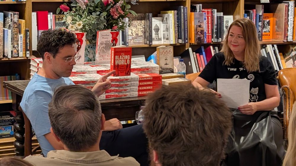 Jon Healey and Rebecca Rideal sit in front of an audience, with bookshelves behind them