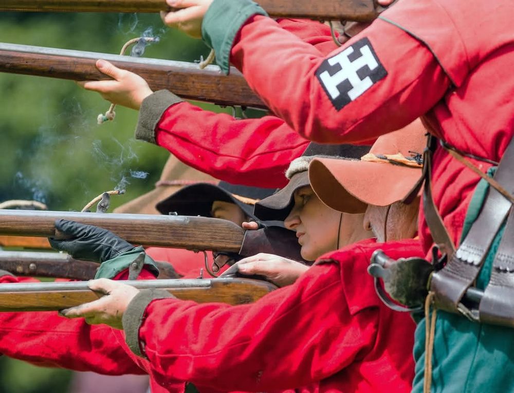 An image of English Civil War reenactors portraying musketeers. They have levelled their muskets at the enemy, wisps of smoke emanating from lit cord held in the mechanism of their muskets. Photo by Laura Johnson 