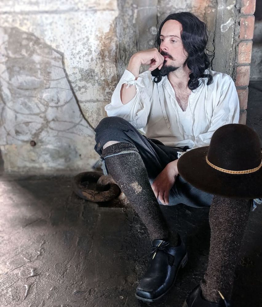 An image of a man in 17th Century clothing – linen shirt, britches, hose, and black leather shoes, with a felt hat balanced on his knee – sits on the floor of a dirty indoor space, leaning back on a wall with broken plaster and exposed brick. Photo by Michael Molcher 