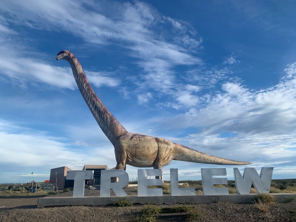 Life sized Patagotitan sauropod statue standing behind a metal sign spelling “Trelew”