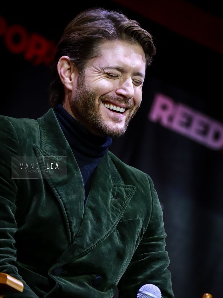 Jensen Ackles sitting on stage in his green velvet smoking jacket and navy turtleneck, holding a microphone in his hand. He is turned a bit to the side and he is laughing so hard his eyes are closed, showing off the gorgeous eye crinkles, and his mouth is open, showing one row of perfect white teeth. His nose is slightly scrunched also.