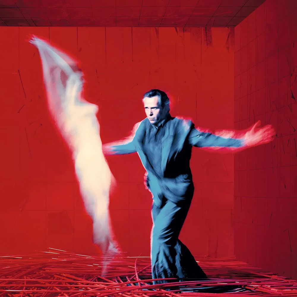 A man in a grey suit stands with arms spread wide against a red background. It's blurry with motion. There's a massive white tear in the shape of a woman to his left. The floor is made of tubes