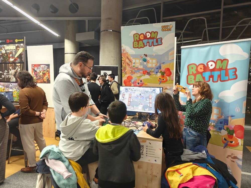 A booth showcasing the video game Roombattle, with family playing on a monitor displaying the game. The background includes a large poster of the game with colorful illustrations.