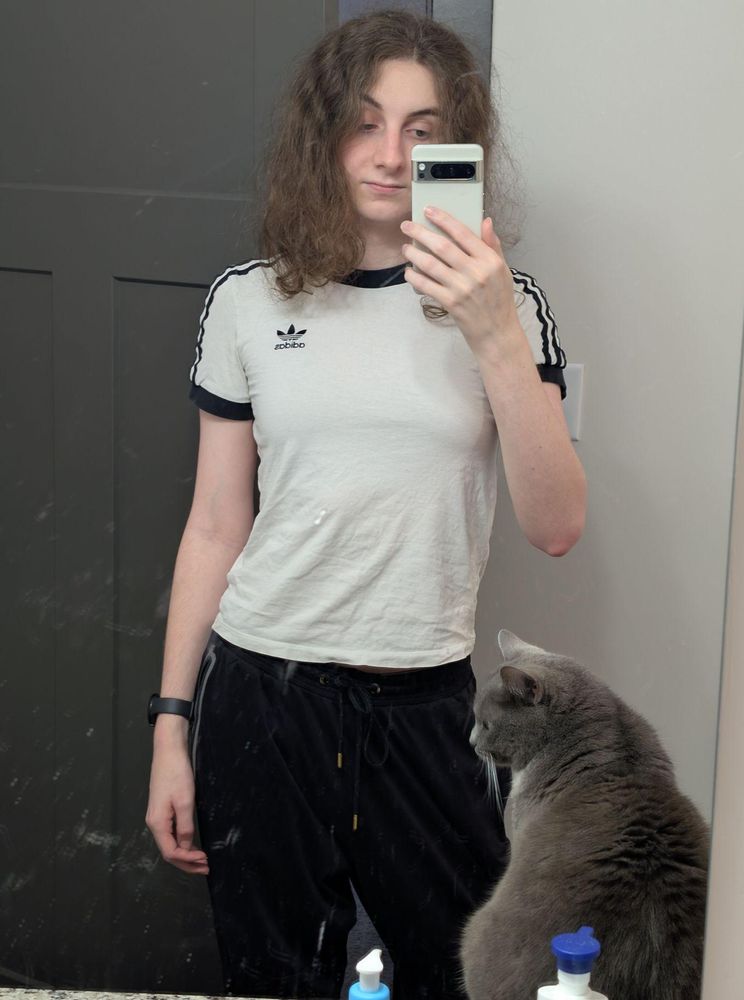 mirror selfie of me in a tight white adidas t shirt and black joggers. my hair is messy. sox the grey and white cat is on the bathroom counter next to me.