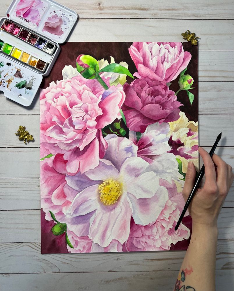 Watercolor painting of a bouquet of flowers. The painting is borderless and fills the page. There are multiple different kinds of flowers mostly in hues of pink. The artists arm and hand are in the picture holding a paint brush. Some of the paints used are also in the image. 