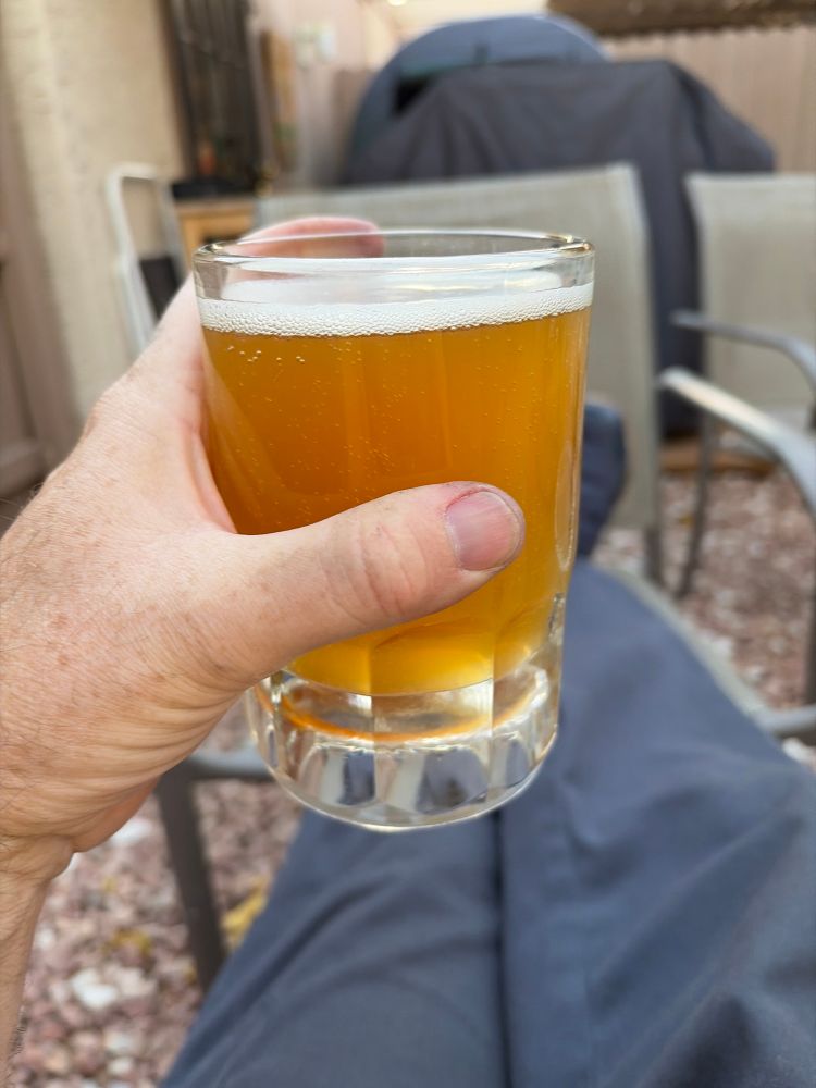 A celebratory golden ale is held up. The drinker’s elevated feet are seen in the background.