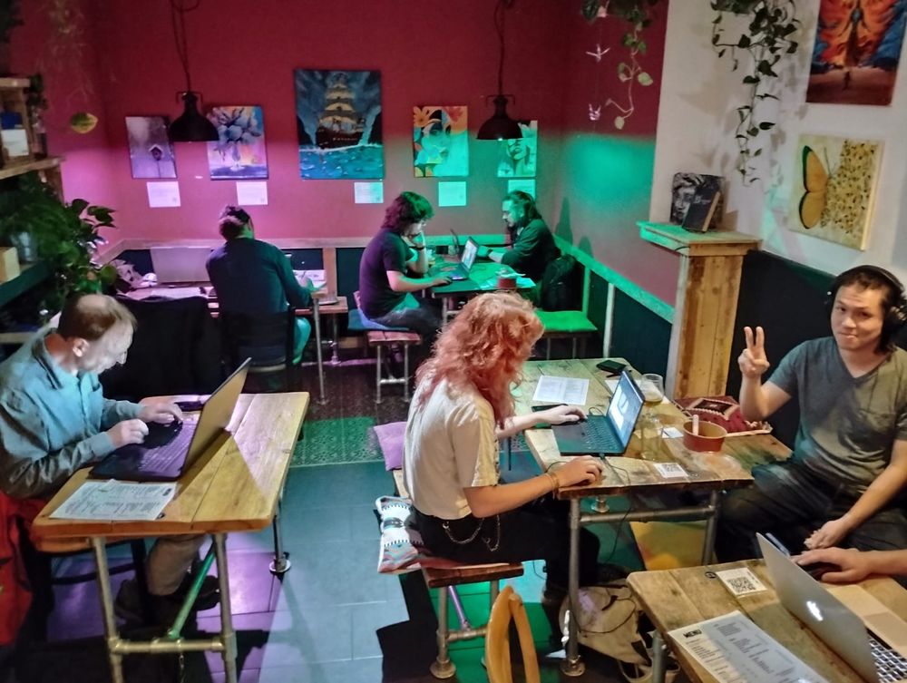 Game developers huddled around a few tables, working on games. Doug is looking at the camera and doing a cheeky peace sign.