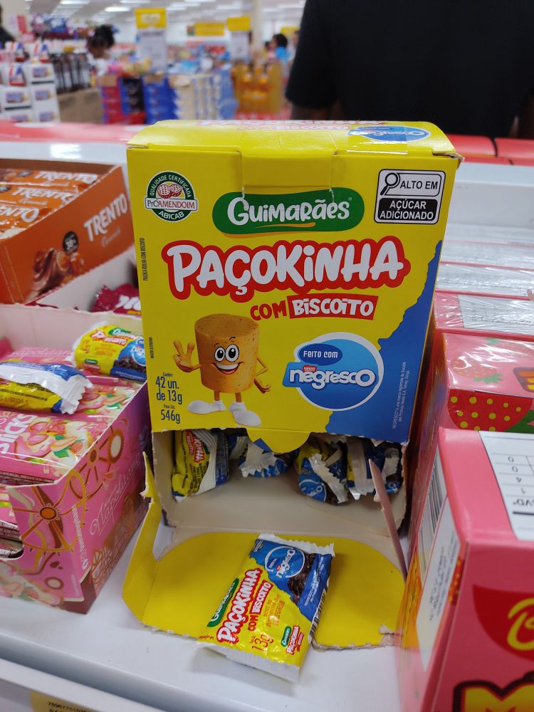Foto da prateleira de um mercado, próximo ao caixa. A foto mostra no centro uma caixa contendo paçocas. As paçocas em questão, são fruto de uma colaboração entre empresas diferentes, no caso, a Paçokinha (da empresa Guimarães) com o biscoito Negresco. Uma combinação que não tive coragem de experimentar, então não poderei descrever o sabor.