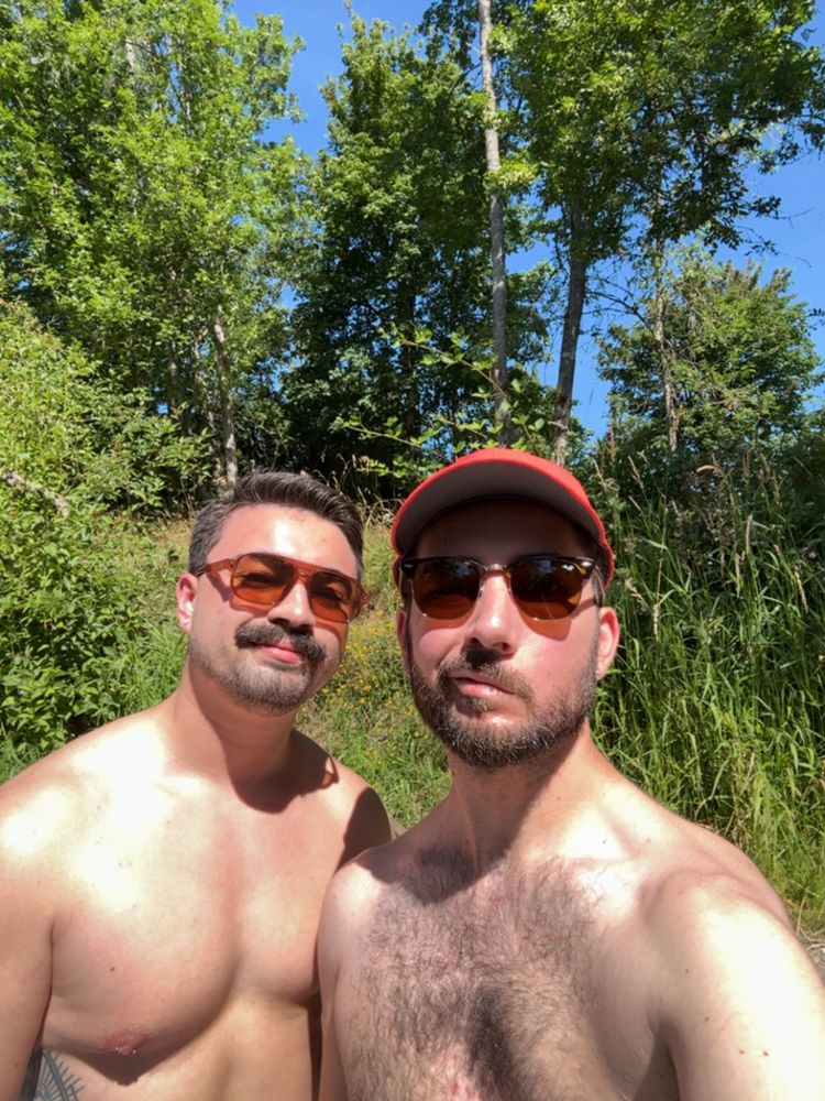 Two shirtless men in sunglasses with greenery behind them. 