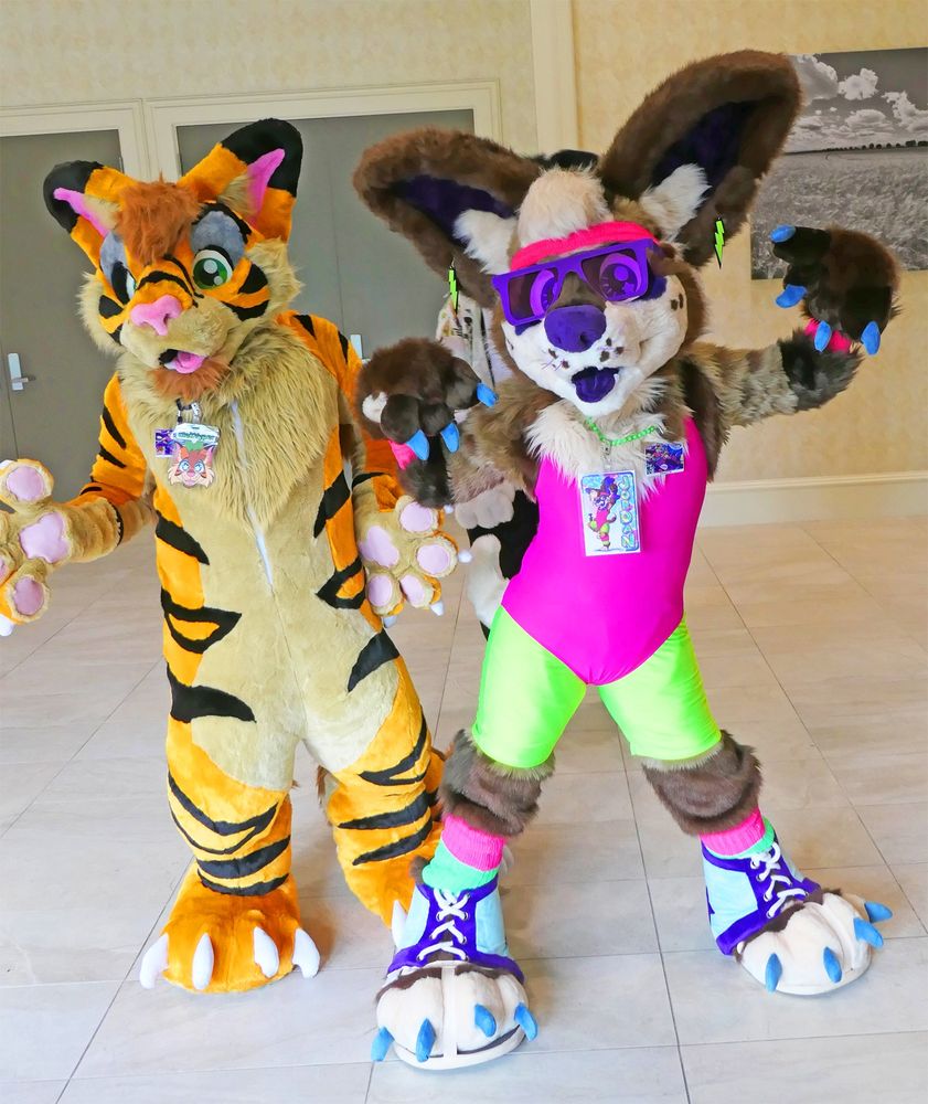 Two fursuiters standing in a lobby of a convention center. On the left is Mek Vyper, a tiger/dragon hybrid. On the right, is Jordan Chihuahua. Mek is facing the viewer with his hand paws extended slightly. Jordan is wearing a stereotypical 1980s aerobics instructor outfit which consists of lightning bolt earrings, a headband, purple sunglasses, a plastic beaded necklace, a one-piece swimsuit, shorts, legwarmers, and fursuit sneakers. Jordan is posed with both arms up in a flex.