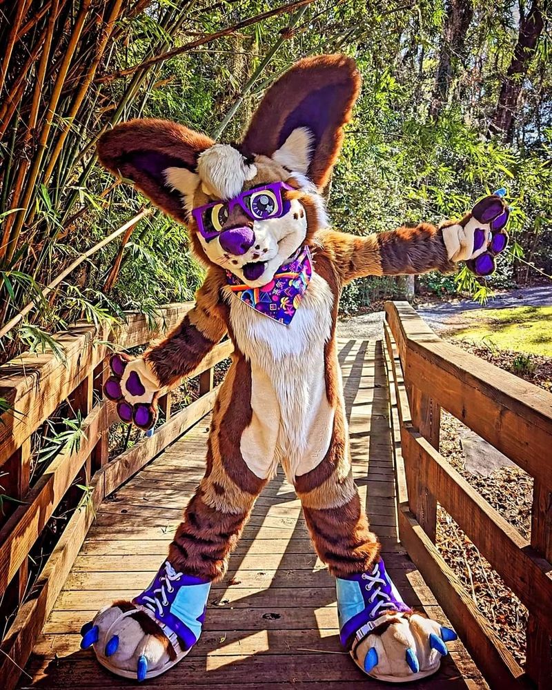 A chihuahua fursuiter is standing on a bridge in a public nature park, facing you, with their arms spread wide for a hug.