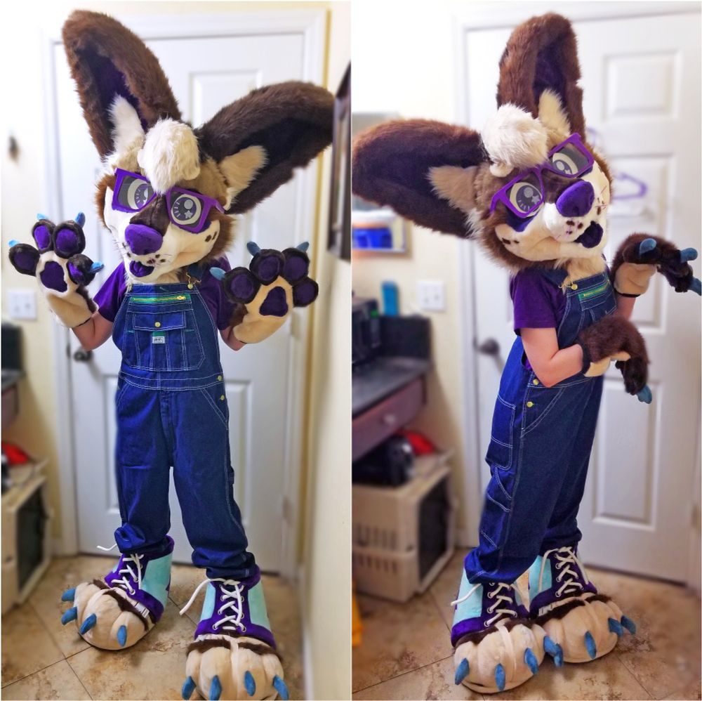 A chihuahua fursuiter is standing in a bathroom wearing overalls and posing cutely for the camera.
