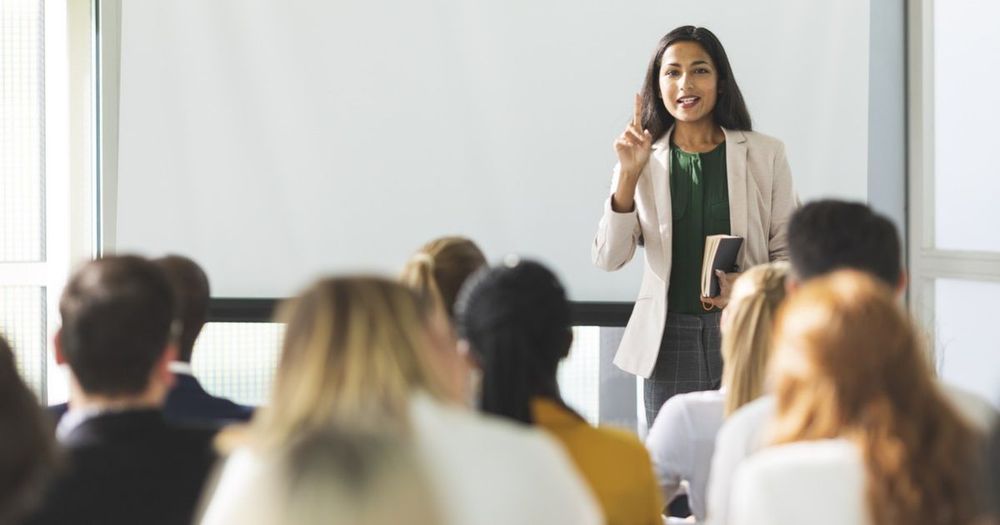 A female speaker stands in front of a classroom, holding a book and gesturing as she addresses students. 