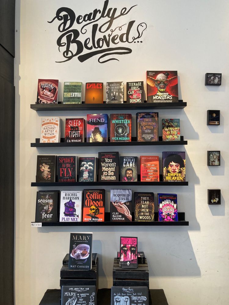 At the Little Ghosts Bookstore, a sign reads DEARLY BELOVED…
Below are the four shelves of new in stock, full of new horror books.
Underneath are the two book club picks.