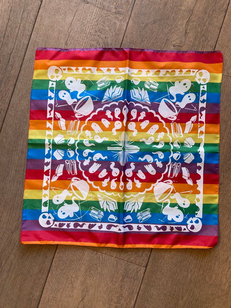 The little ghosts rainbow bandanna! Depicts a pattern of our logo with a banner of skulls and ghosts, open books inside, and an interior pattern of a mixture of wiggly and sheet ghosts.