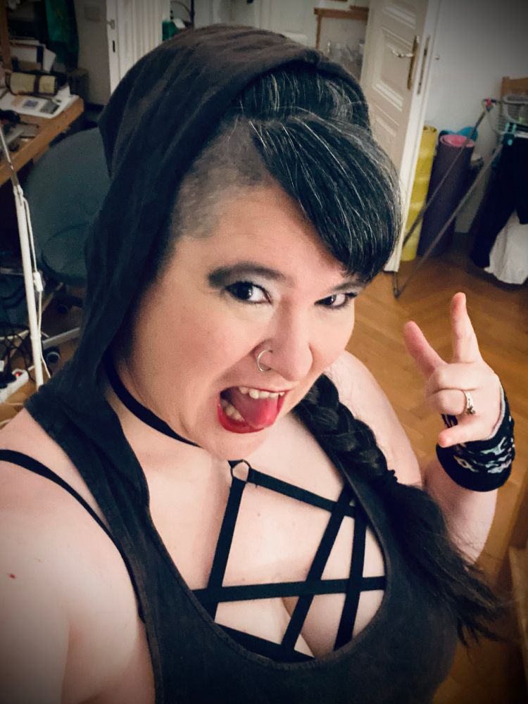 Jen wearing a sleeveless top with a hood and a pentagram under it. She has shaved sides and a side bang and very 2000s smoky make up and is making a horn sign 