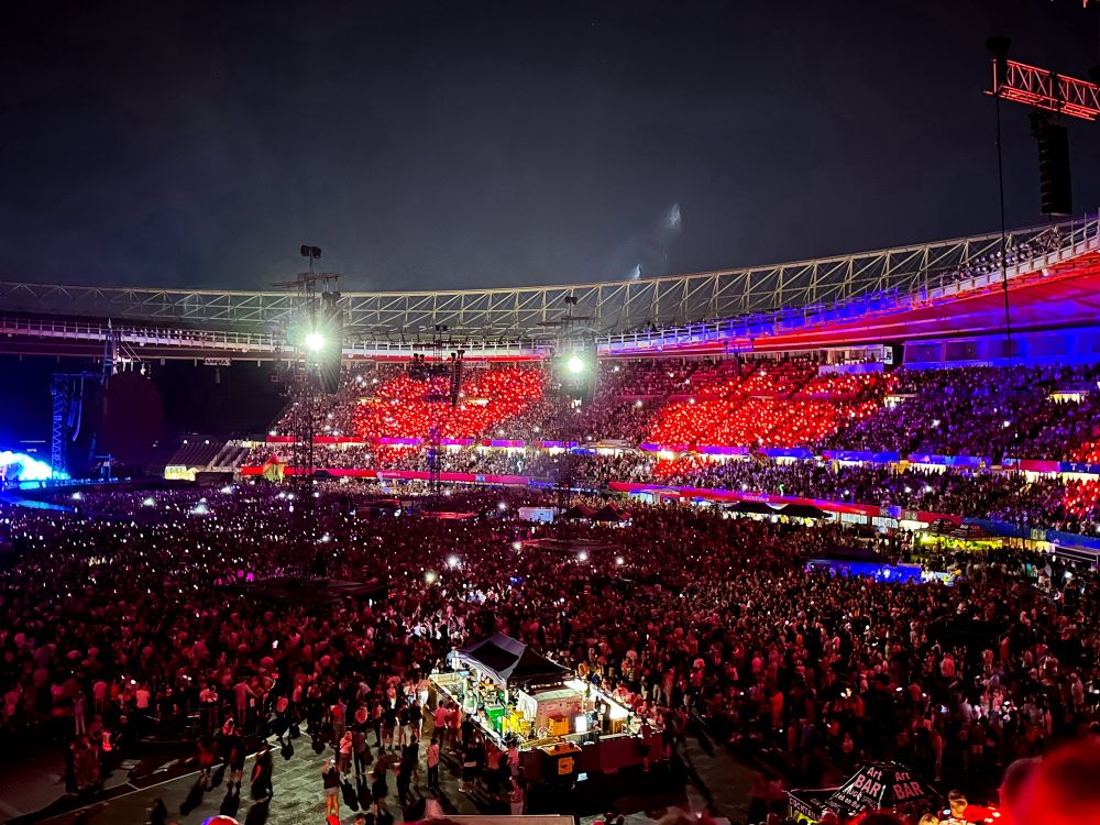 View on the concert scene from the seats on the upper rows. Some kind of red era as there are two hearts seen on the opposite side of the stadium cause the wristbands people had on there arm were lit red and formed these  hearts.
