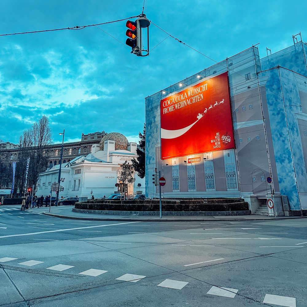 A big billboard advertising from Coca-Cola hanging on the side of a well wrapped building. Lights are shining on it. 
The Secession with its golden roof is in the background. The street in front is luckily empty. The Traffic lights are on red yellow. The picture is in portrait format and the filter is highlighting the colours very bright with little blue flair. 