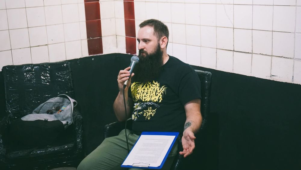 A man in his 30s, long dark beard, sitting on a black, foil-wrapped chair, talking into a mic. Some notes on his lap. Black t-shirt with a metal "Flail to the face" writing and art.