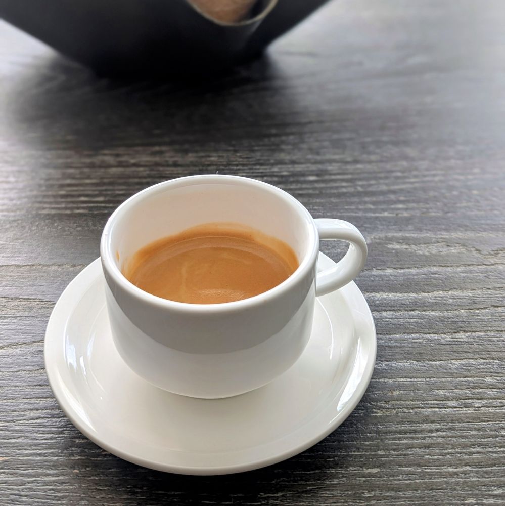An espresso poured in a white cup on a white plate sitting on a grey wooden table. The caffeine is necessary.