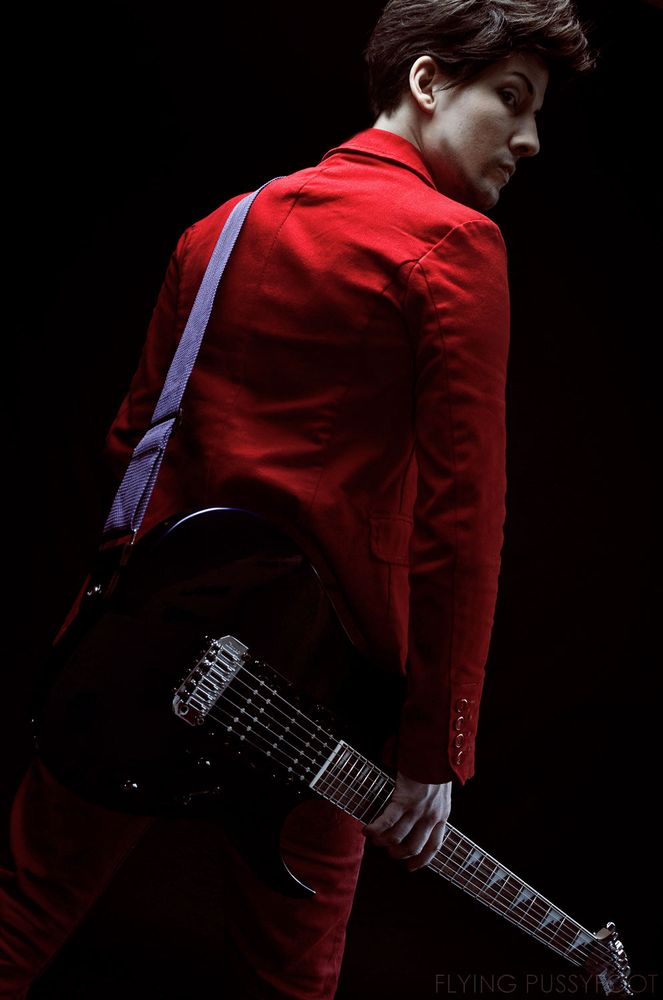 A cosplayer as Matthew Bellamy from the British band Muse in his red suit holding a guitar.