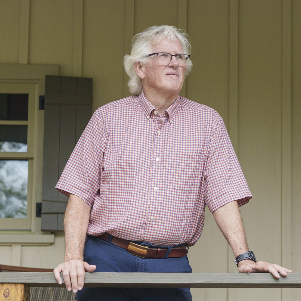 APS President Robert Hester, a white man with white hair and glasses, wears jeans and a checked shirt and is leaning on a wooden railing.