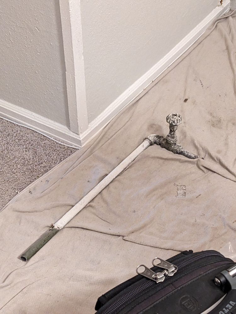 A severed and capped l-shaped metal pipe with a decaying old valve on it. It is painted white, but a gas leak has corroded the fitting somewhat so it has black showing around the cap. It lies on a blanket on the floor, near the hole in a white wall from which it was cut.