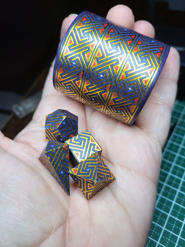 A roll of wide washi tape with an angular gold, red, and blue celtic maze like pattern in the hand of a dice maker. She is also holding four polyhedral shape dice wrapped with the same washi tape.
